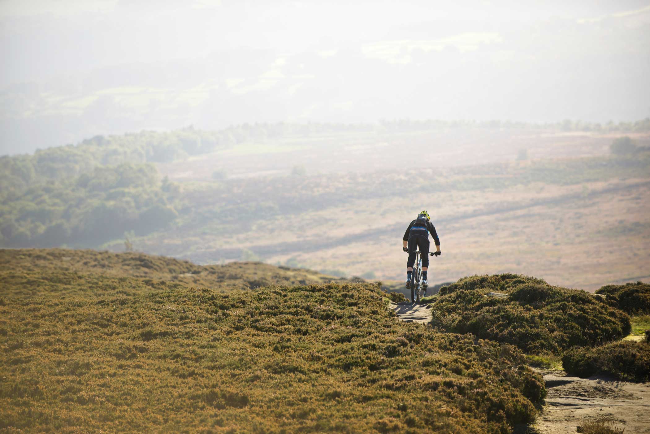 Mountainbikecyklist i svart klädsel cyklar på en stenig stig genom ett kuperat, buskigt landskap.